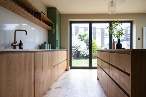 Farblich abgesetzt steht der große Schrank in Grün im Raum und bietet viel Platz für Zubehör. Die Küchenzeile selbst wird durch ein Wandregal aufgelockert, das verleiht dem Raum Leichtigkeit. Alles von Wood Works Brighton.